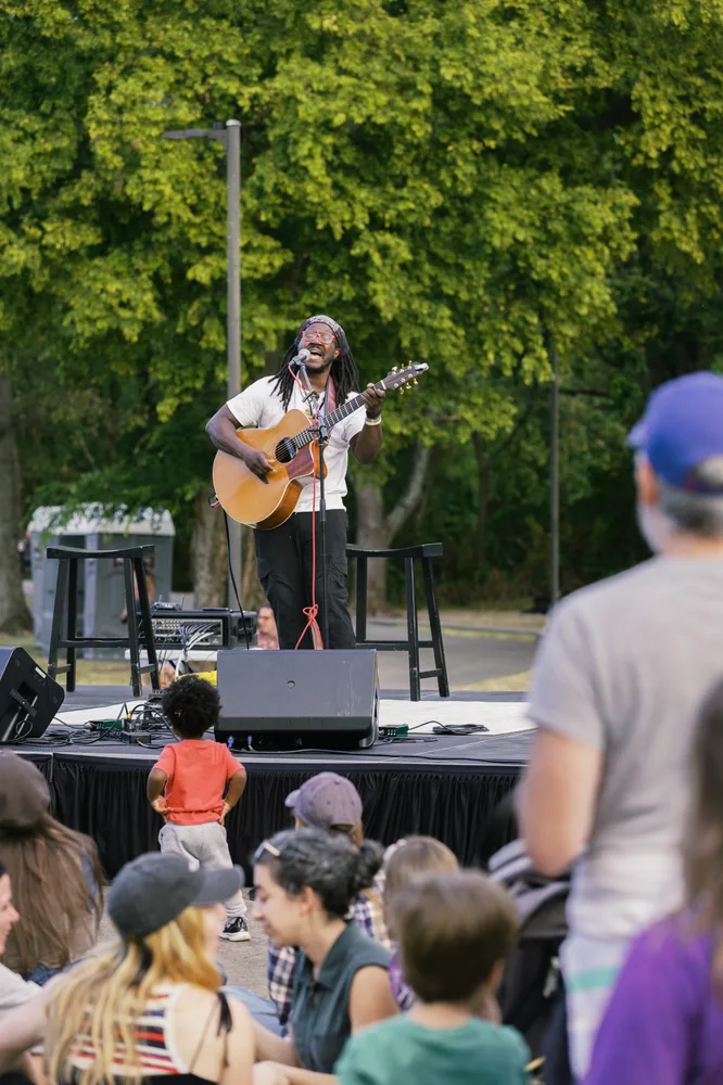 A musician playing guitar on stage for neighbors seated on the lawn.